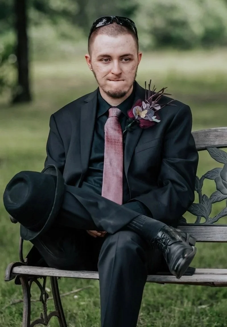 A young man in a black suit with a pink tie and boutonniere, sitting on a wooden bench in a park, holding a black hat in his lap, with sunglasses on his head and a contemplative expression.