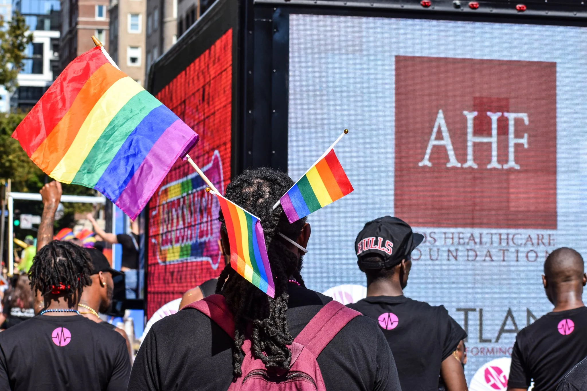 A crowd at a pride parade, with rainbow flags and people wearing black shirts with pink logos. In the background, a large digital screen displays the words 'AHE' and information about a healthcare foundation.