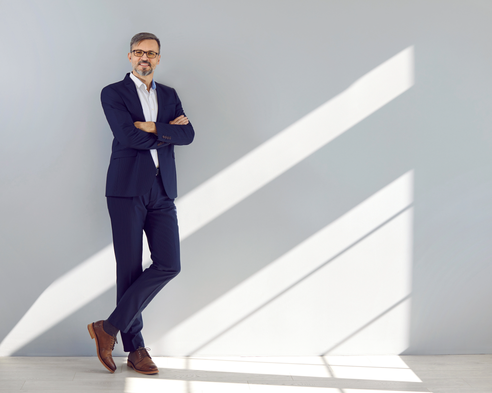A confident man in a navy suit and glasses standing with arms crossed against a light gray wall with diagonal sunlight shadows.