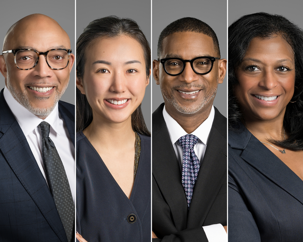 Four professionally dressed individuals posing for a group photo against a gray background.