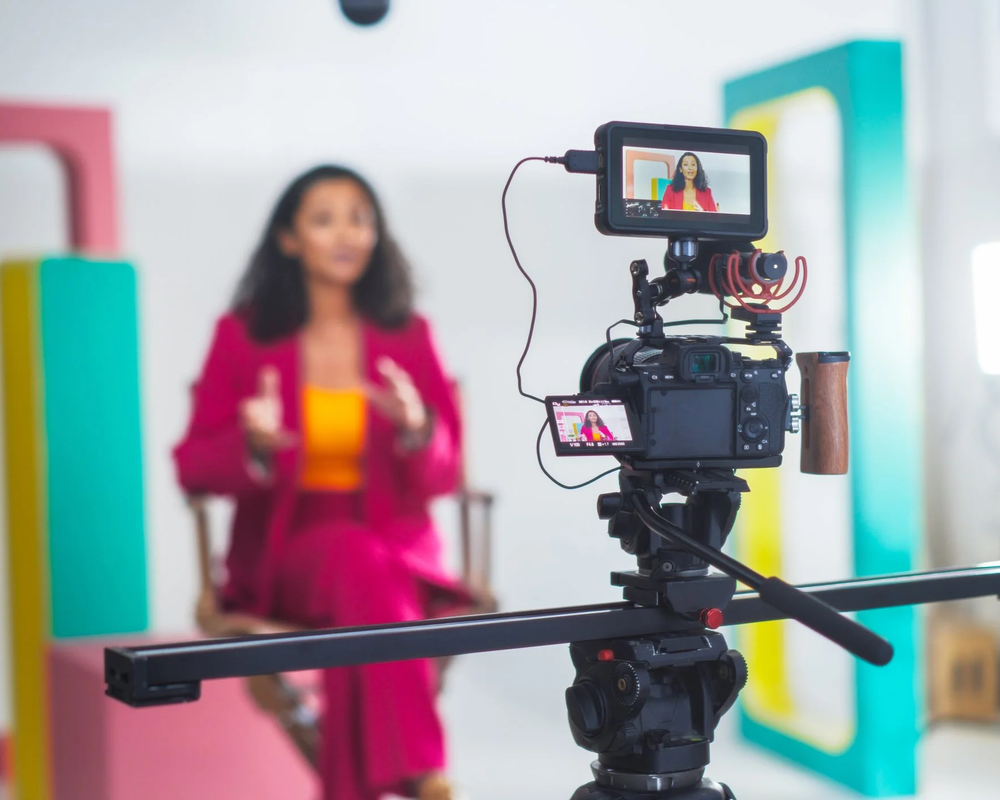 A professional camera setup is recording a woman in a pink suit, sitting in a chair and speaking during a video shoot or interview.