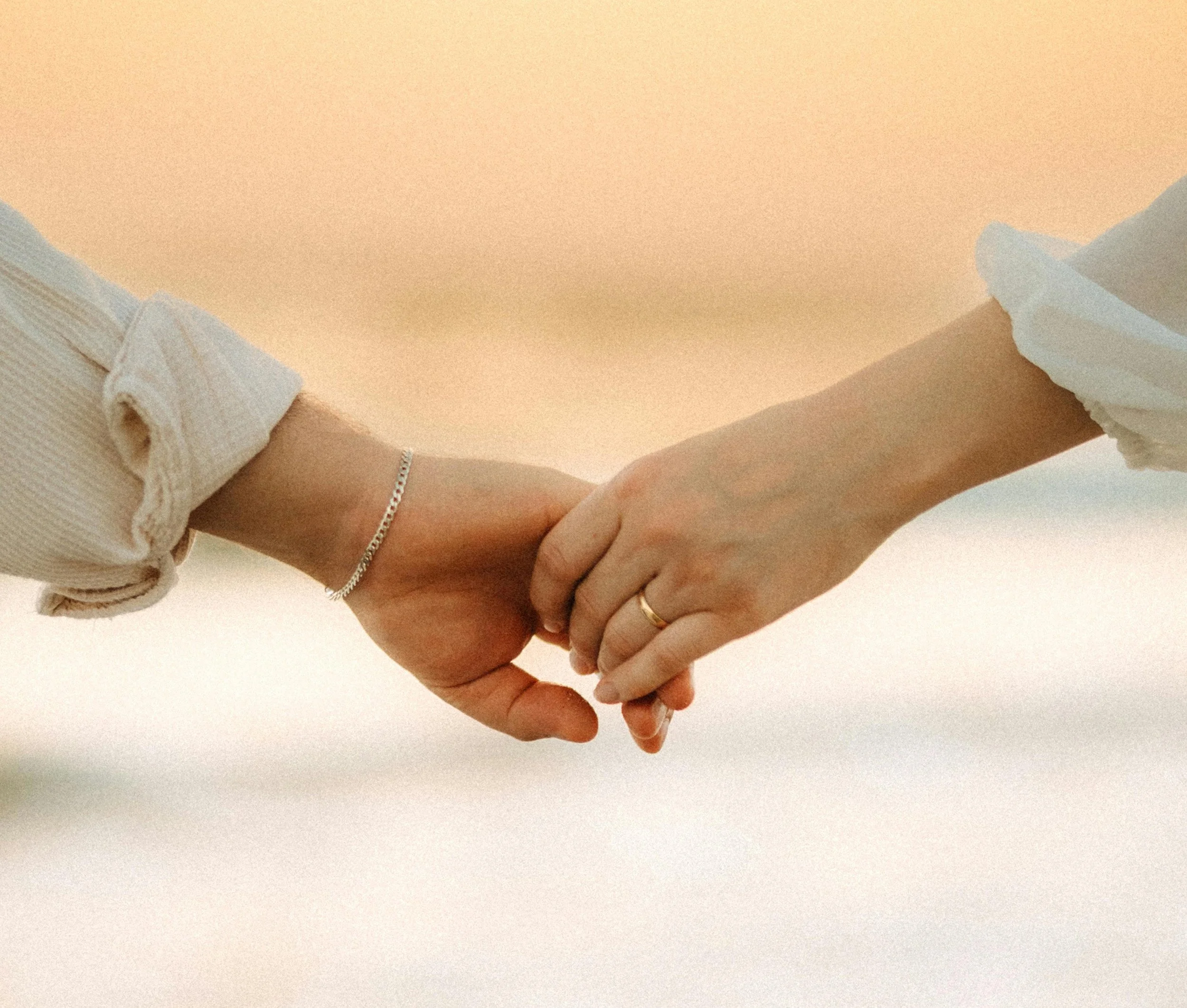 Close-up of two hands gently holding each other, with one hand wearing a ring and a delicate bracelet, against a plain light background.