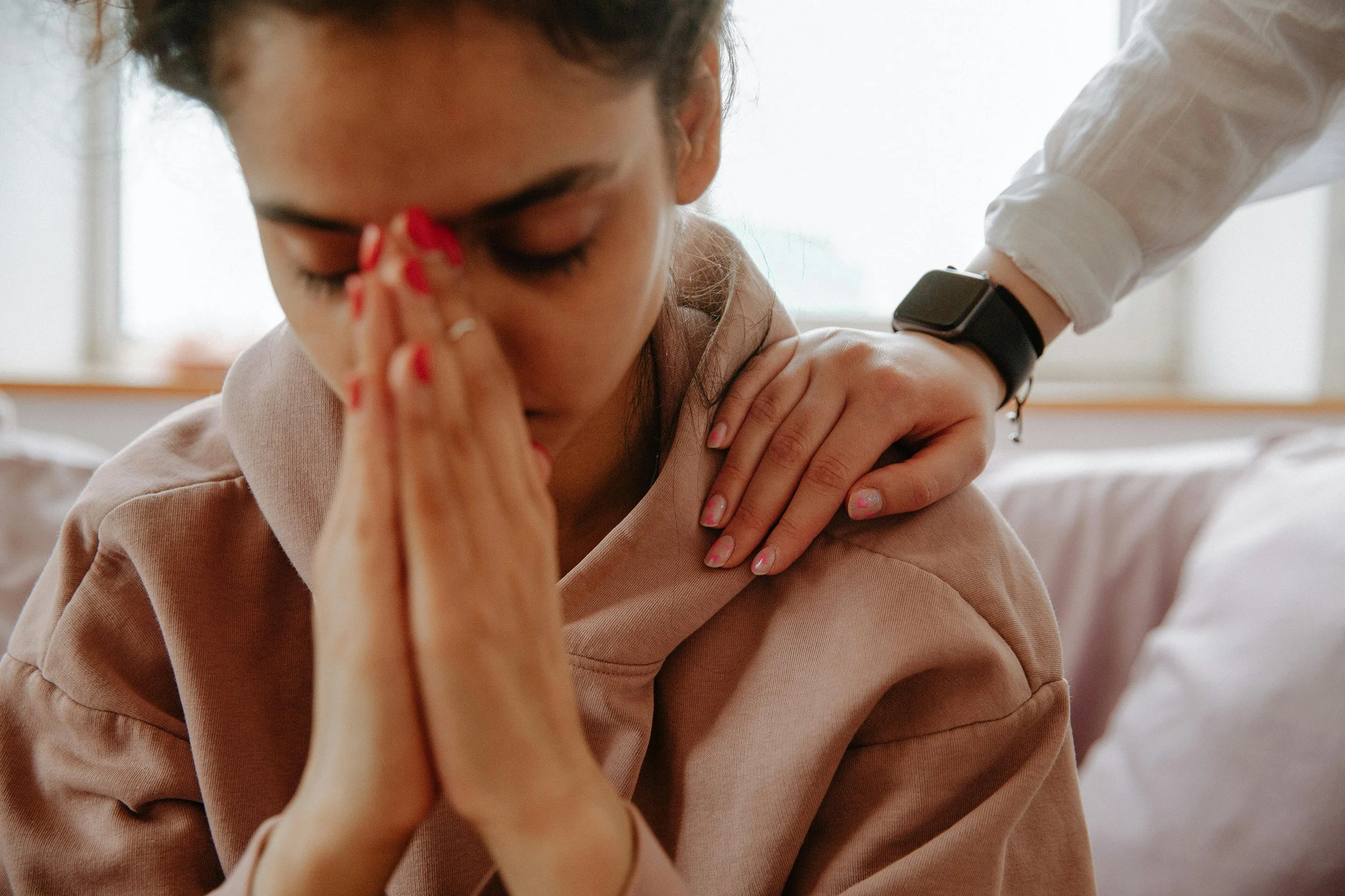 Woman praying with hands together while another person offers comfort by placing a hand on her shoulder.
