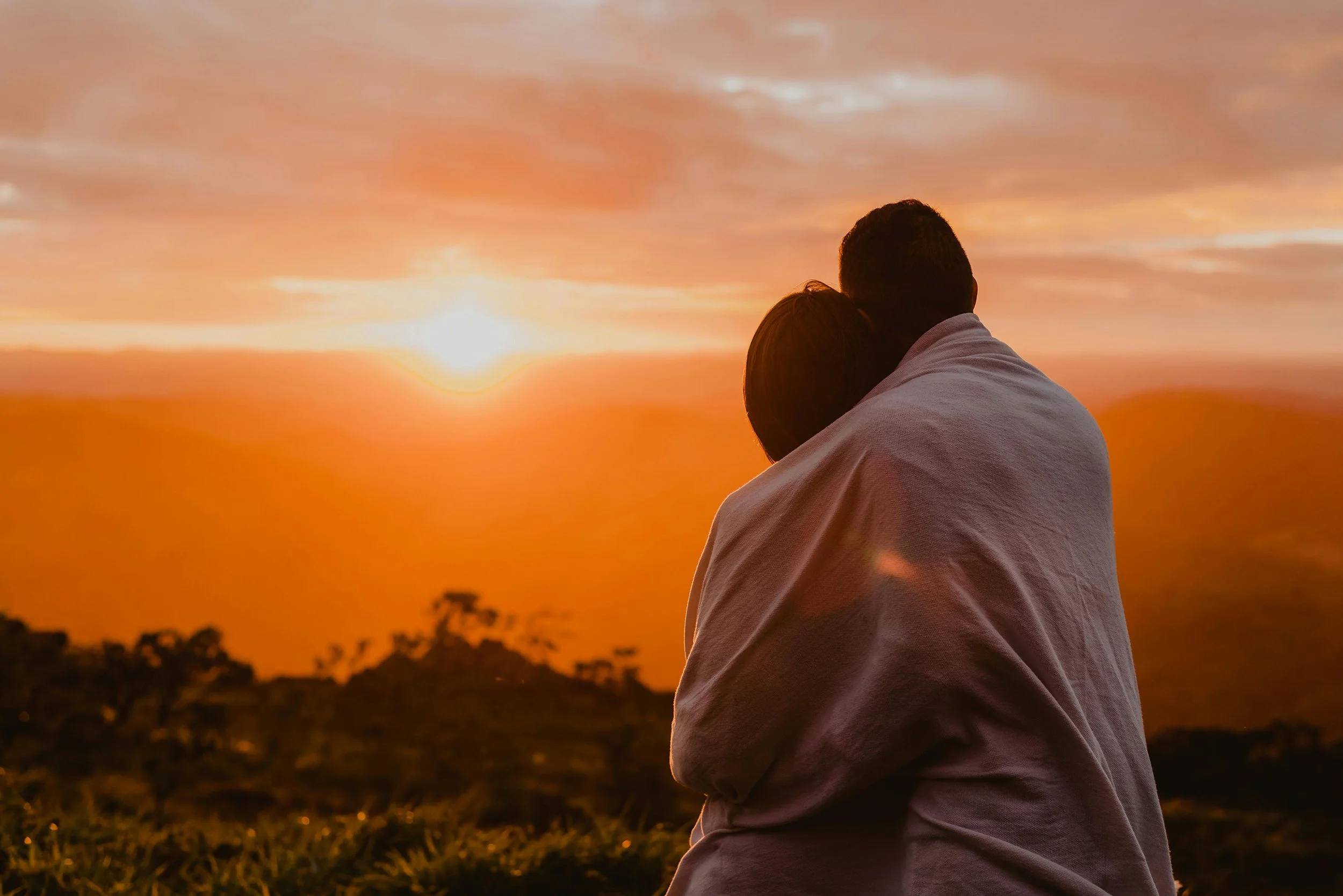 A couple wrapped in a blanket watching a peaceful sunset together over a mountain landscape.
