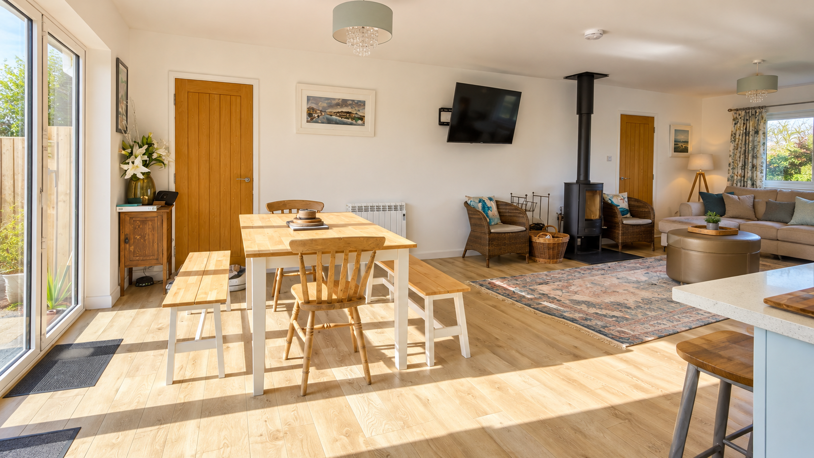 Open-plan living and dining area with wooden flooring, a dining table with benches and chairs, a sofa with cushions, a coffee table, a fireplace with rattan chairs, a wall-mounted TV, and large windows with curtains letting in natural light.