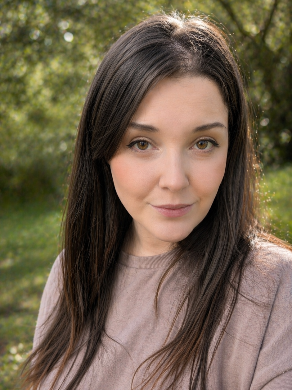 A young woman with long brown hair and brown eyes posing outdoors with greenery in the background.