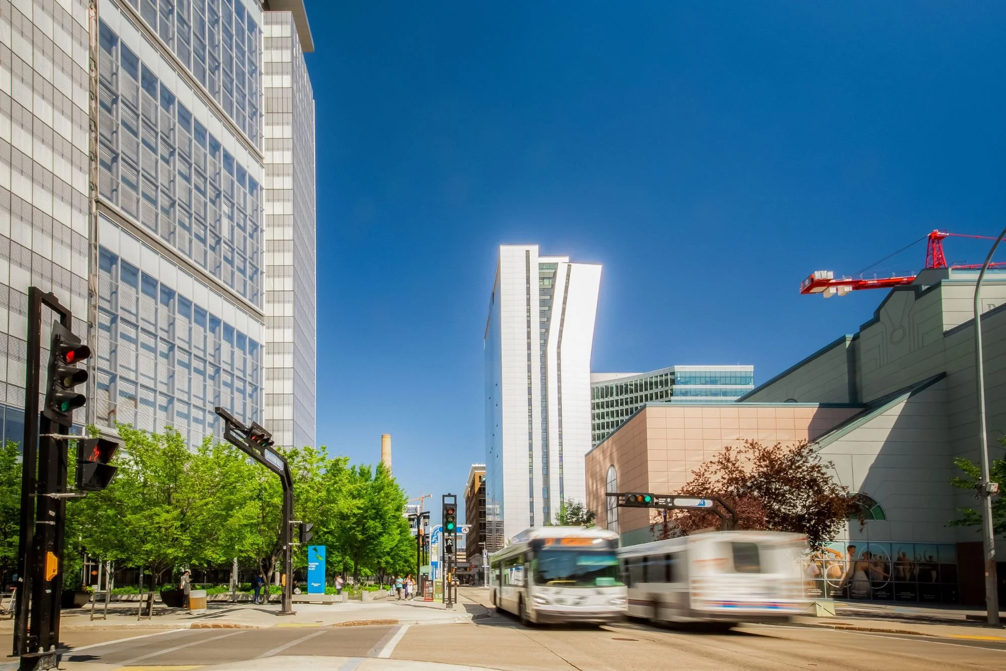 A busy city street scene with tall modern buildings under a clear blue sky. A bus is moving in the foreground, and traffic signals are visible at the intersection. There are street signs, greenery, and a few pedestrians on the sidewalk.