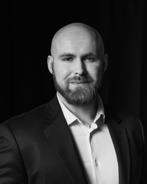 Black and white headshot of a man with a bald head and beard, wearing a suit jacket and collared shirt, looking confidently at the camera.