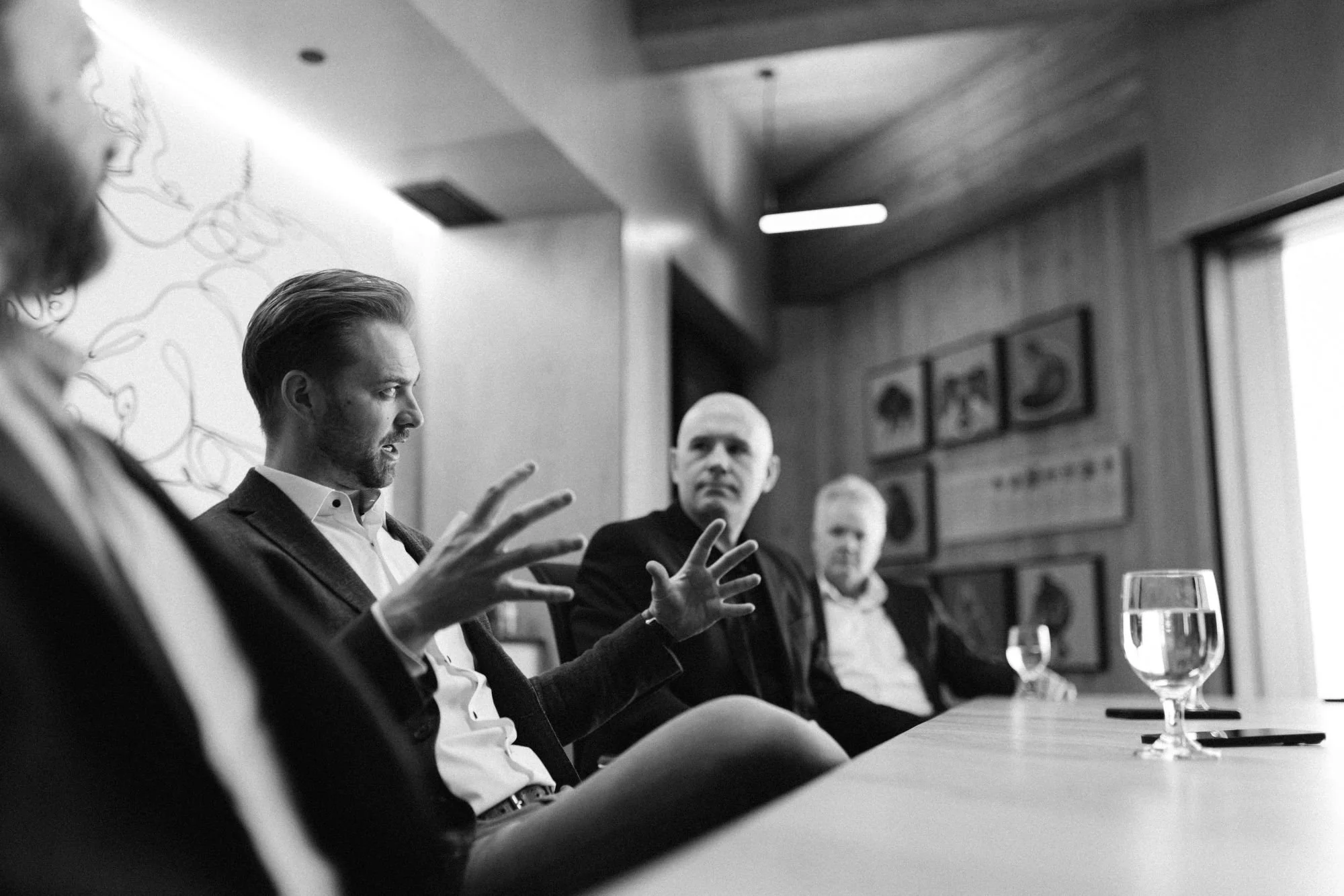 A black and white photo of a man speaking at a business meeting, seated at a conference table with other attendees. The man is gesturing with his hands. The setting appears to be a formal office or conference room.