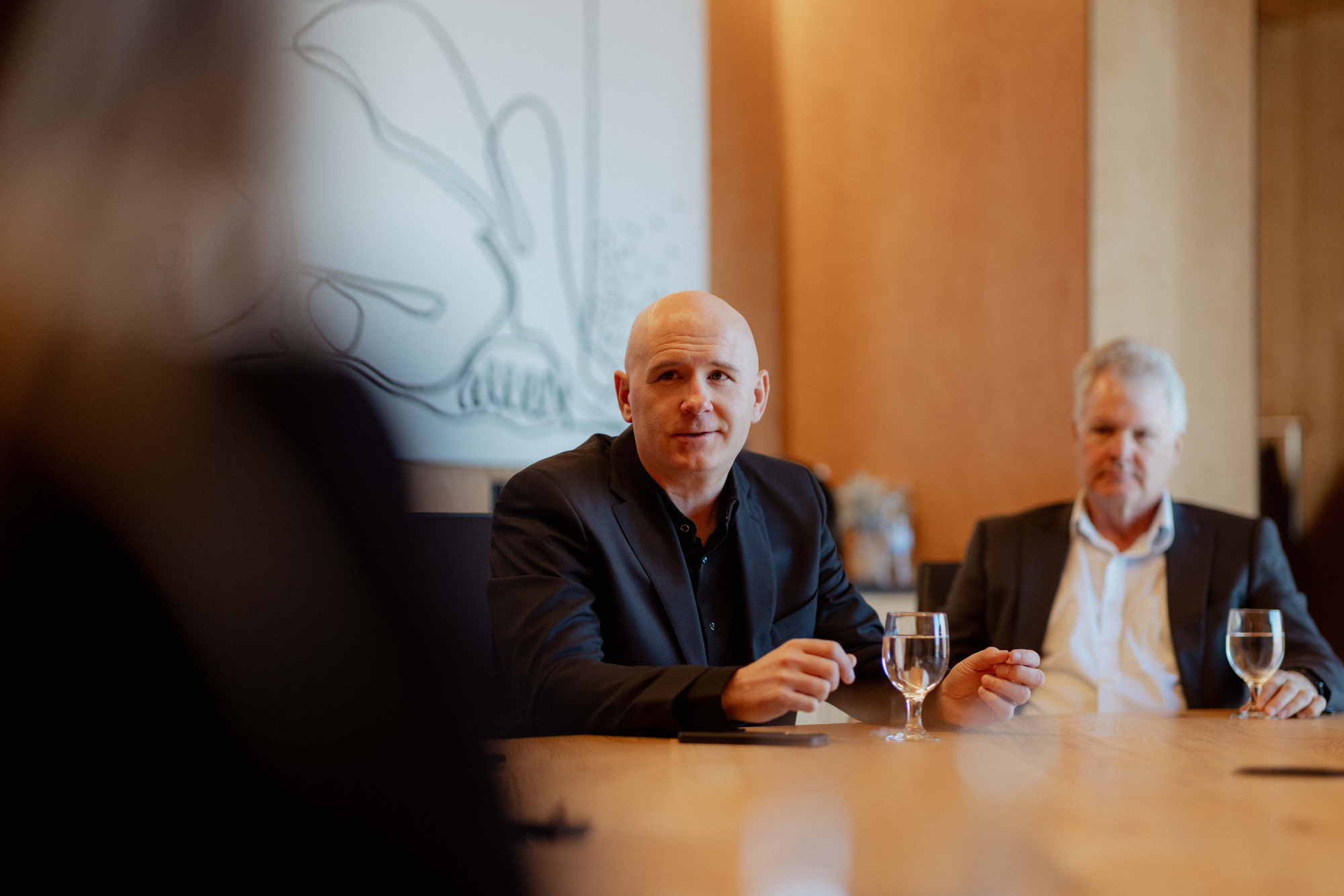 Two men sitting at a conference table with glasses of water, engaged in a discussion. One man is bald and wearing a black suit, while the other has gray hair and is wearing a white shirt and dark jacket. The scene takes place in a room with wooden panels and a whiteboard or projection on the wall.