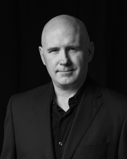 Black and white portrait of a bald man wearing a dark suit jacket and shirt, looking at the camera with a slight smile.