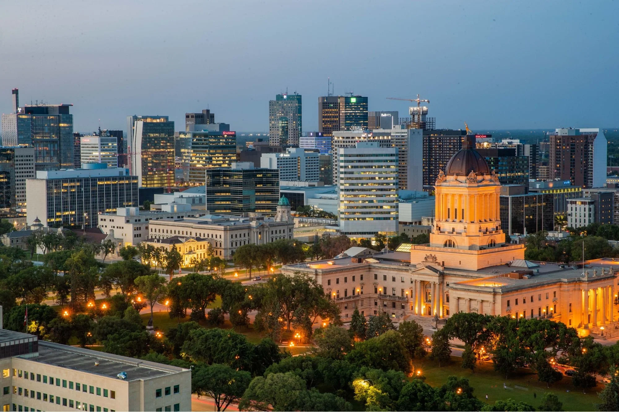 City skyline at dusk with illuminated office buildings, a historic government building with a dome, and trees in the foreground.