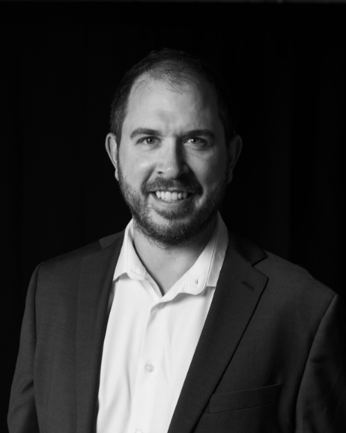 Black and white portrait of a smiling man with short dark hair and a beard, wearing a suit jacket and white shirt against a dark background.