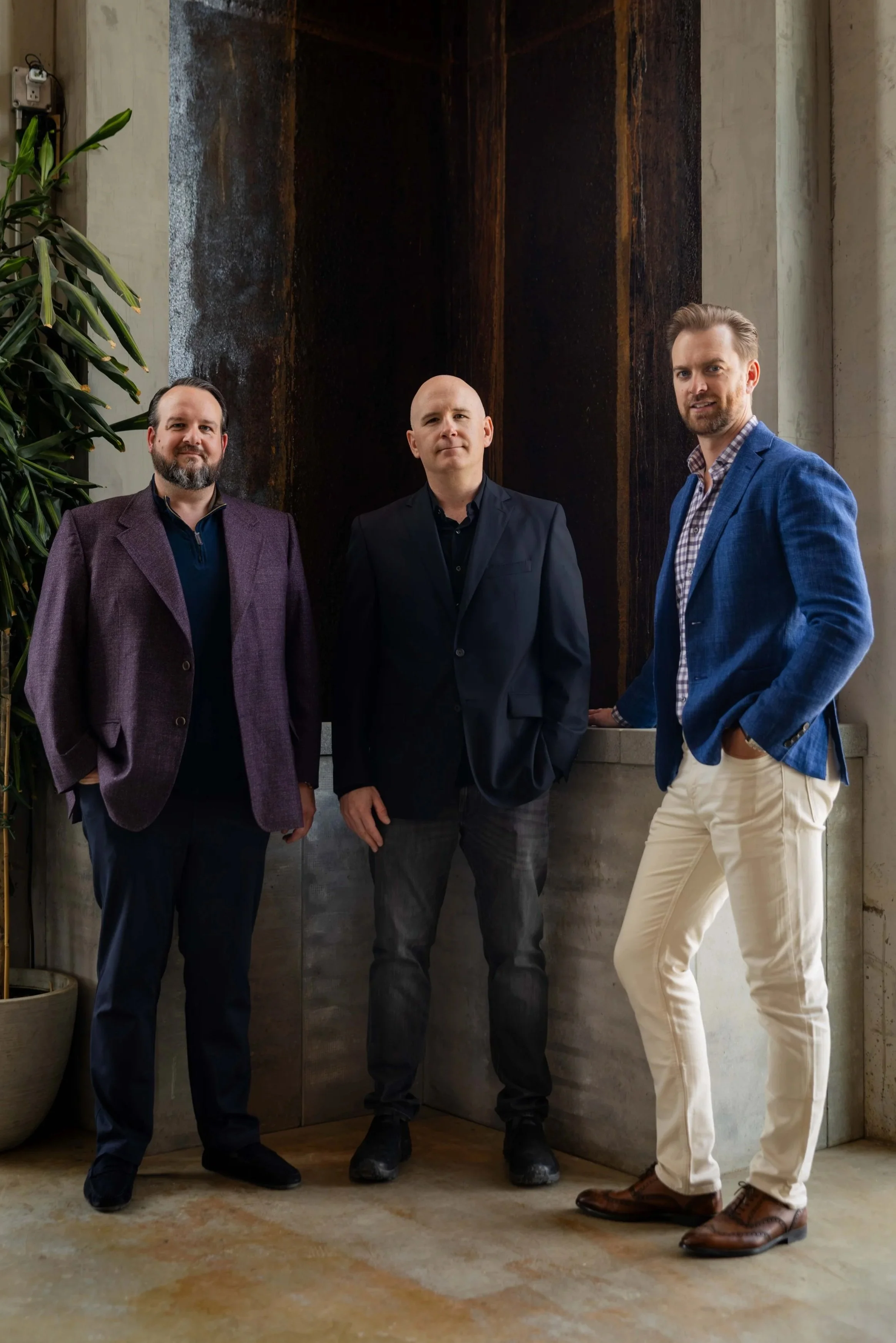 Three men standing indoors in front of a dark wall with a plant on the left. They are dressed in smart casual and formal attire, looking at the camera.