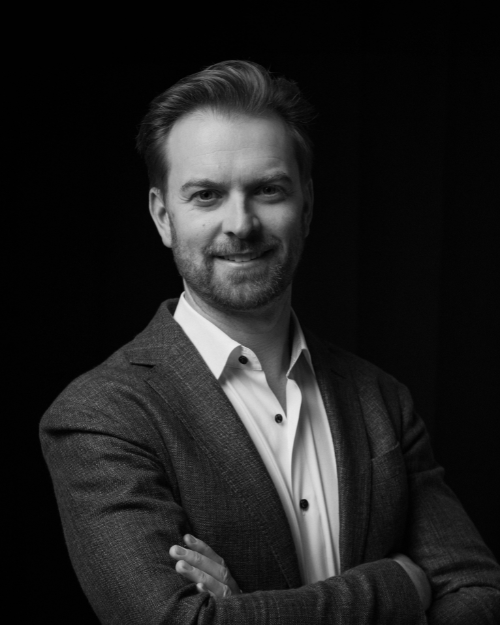 Black and white portrait of a man in a blazer with arms crossed, smiling at the camera.