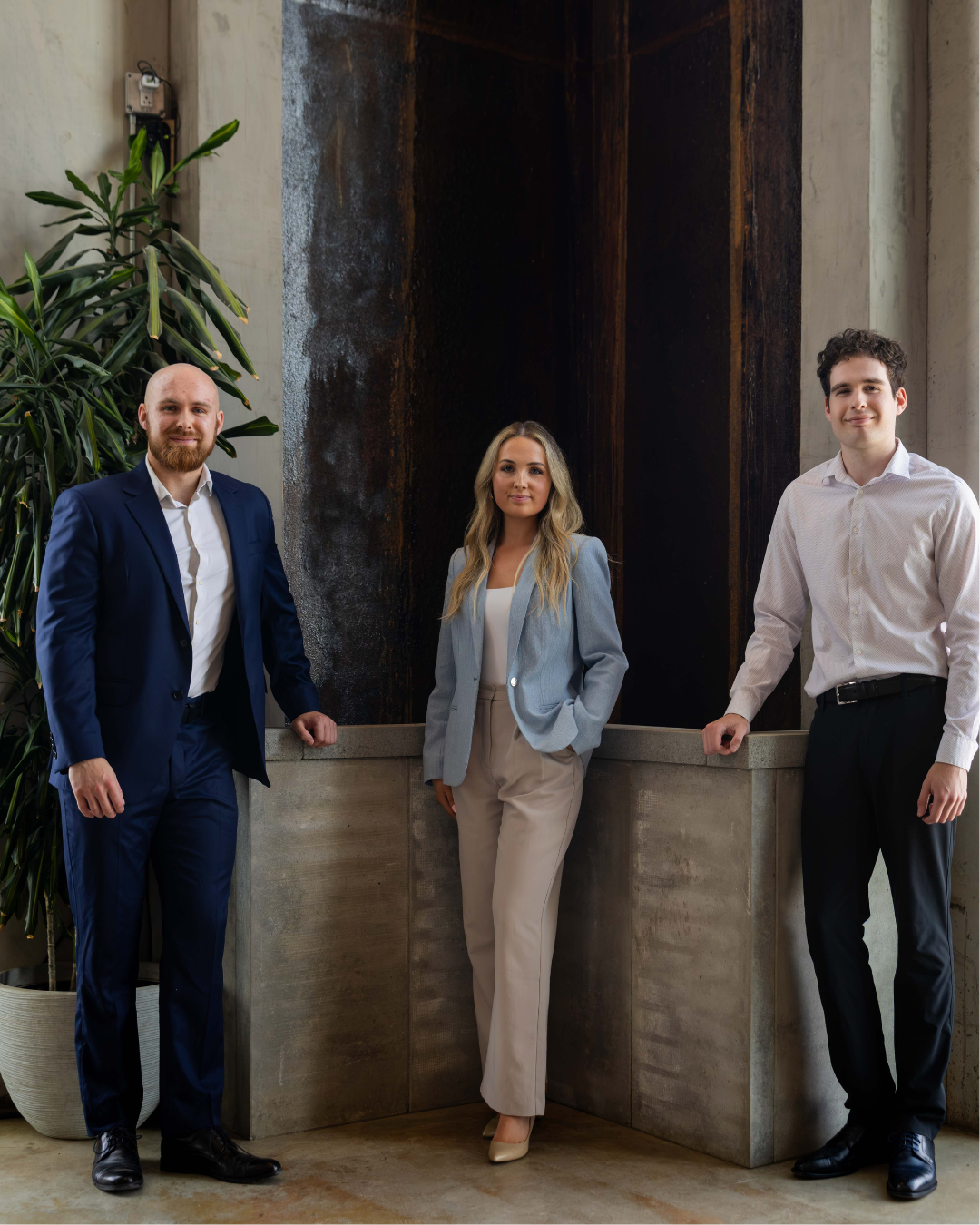 Three professionally dressed individuals standing indoors in front of a modern wall with greenery on the left side.
