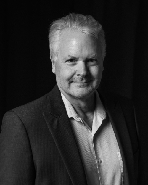 Black and white portrait of a smiling older man with gray hair, wearing a suit jacket and a collared shirt, standing in front of a dark background.