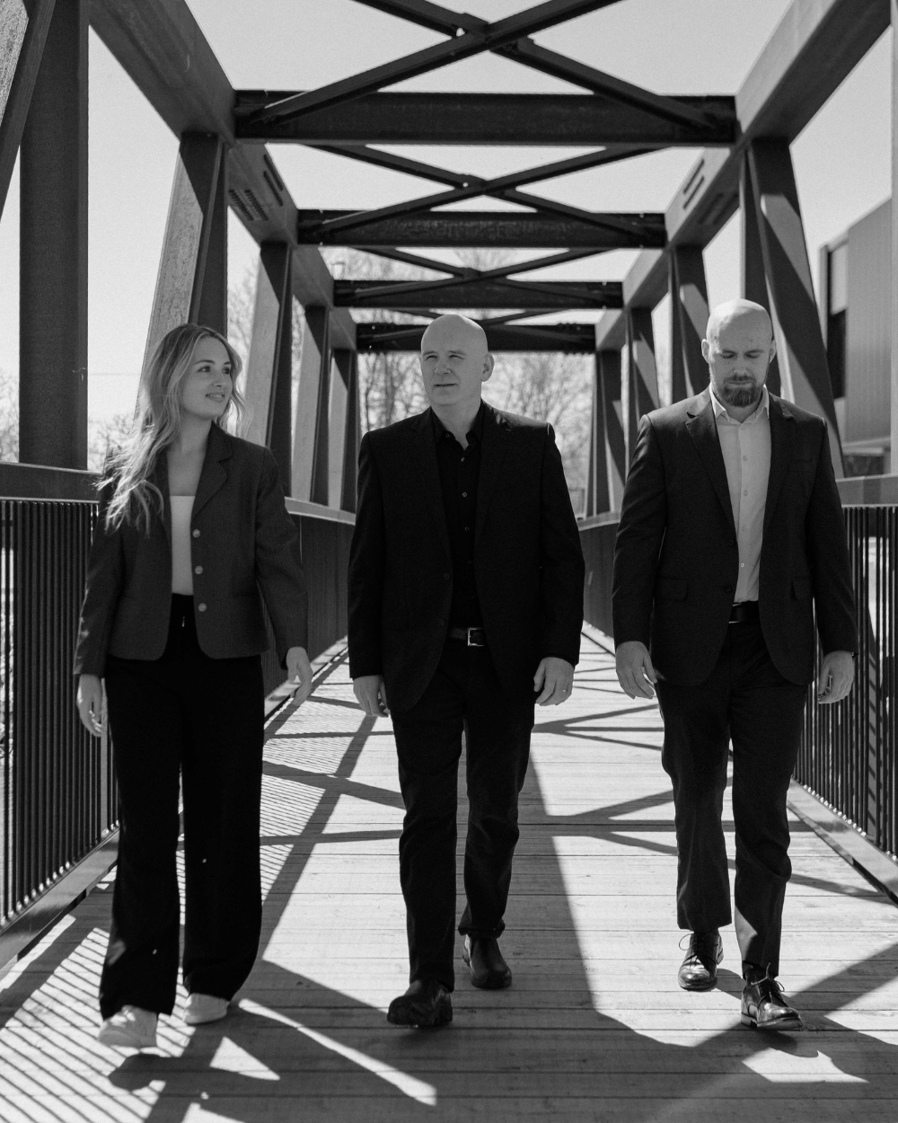 Three professionals, two men and one woman, walking and talking on a wooden bridge with metal railings on both sides, under a metal structure overhead, in black and white.