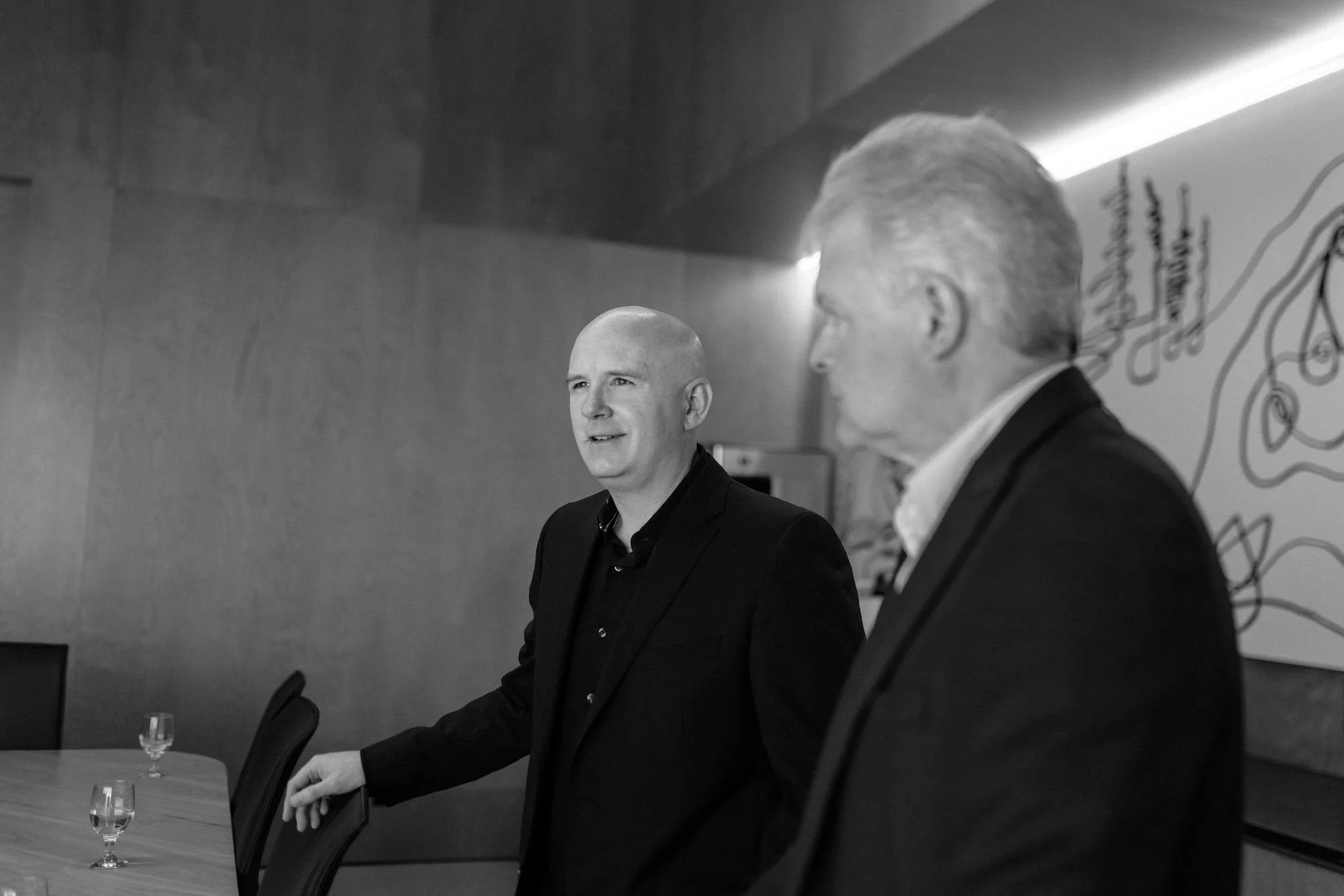 Two men in formal suits standing at a conference table with glasses of water, engaging in discussion, with a decorative wall and artwork in the background.