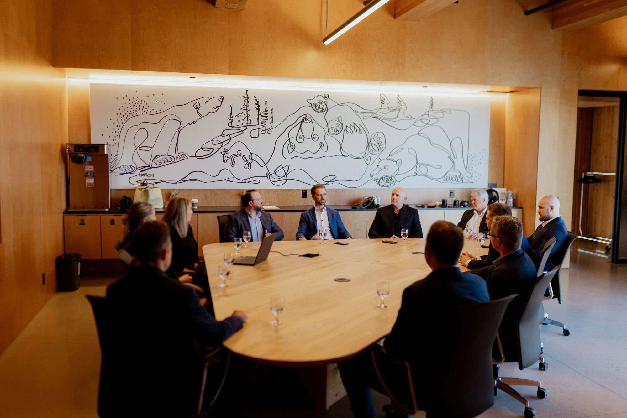 Business meeting in a modern conference room with a large wooden table, around which several men and women in formal attire are seated, facing a wall with abstract line art painting of animals and nature, with natural and ambient lighting.