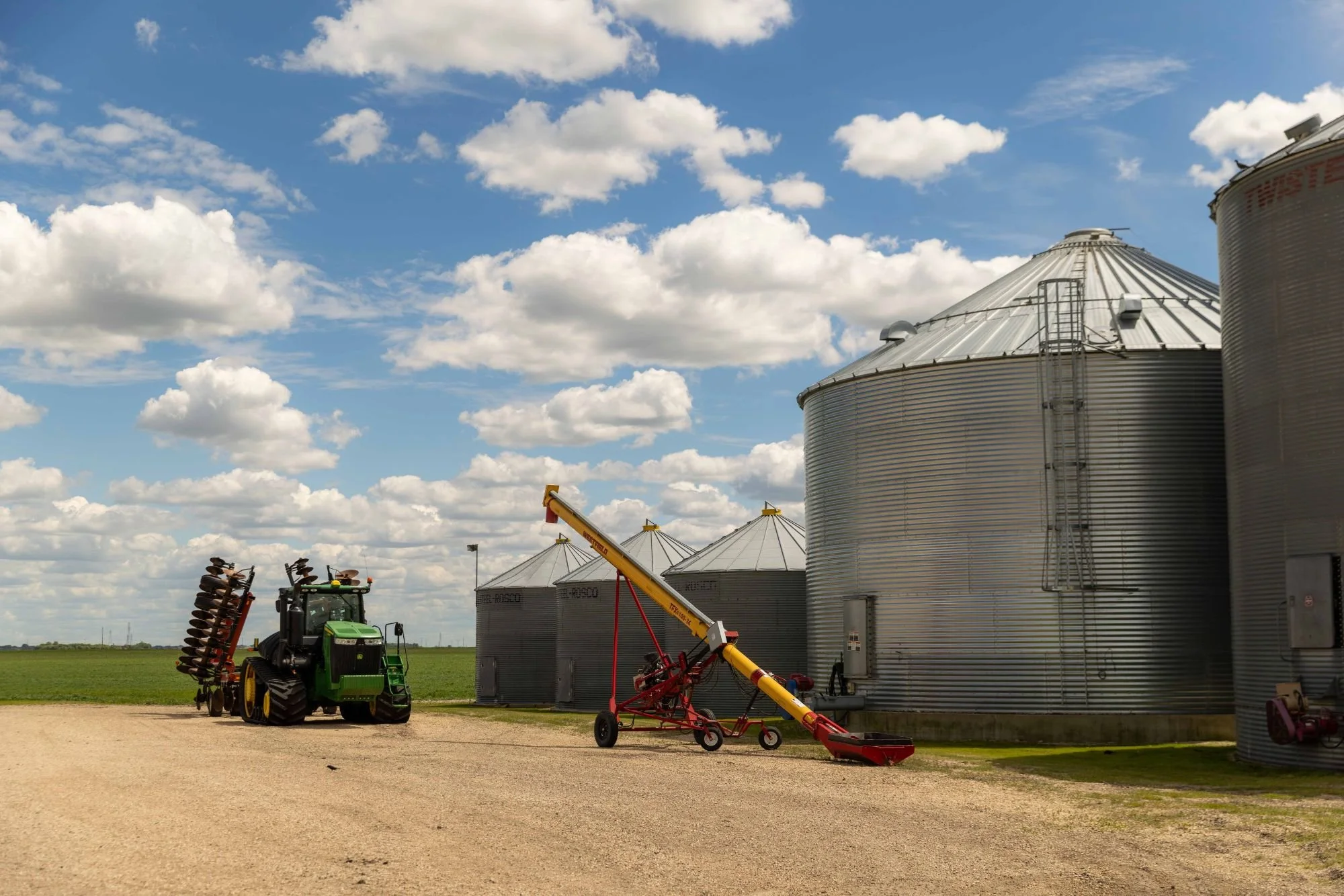 Agricultural equipment next to large metal grain silos under a partly cloudy sky.