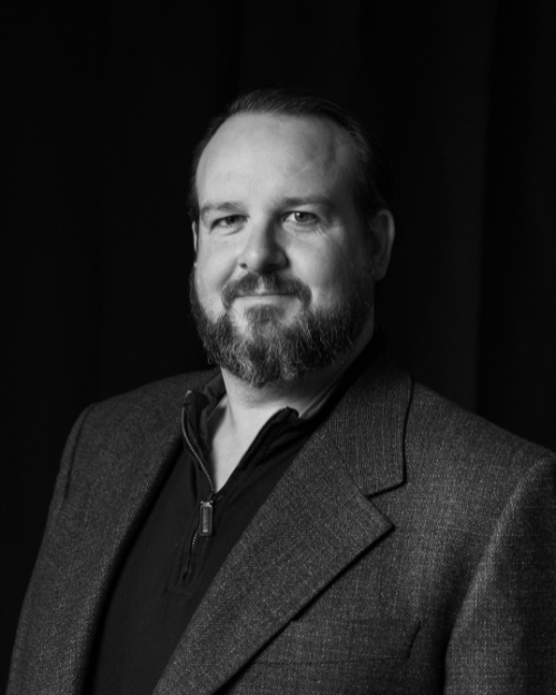 Black and white portrait of a man with a beard, wearing a dark blazer and a zippered shirt, standing against a dark background.