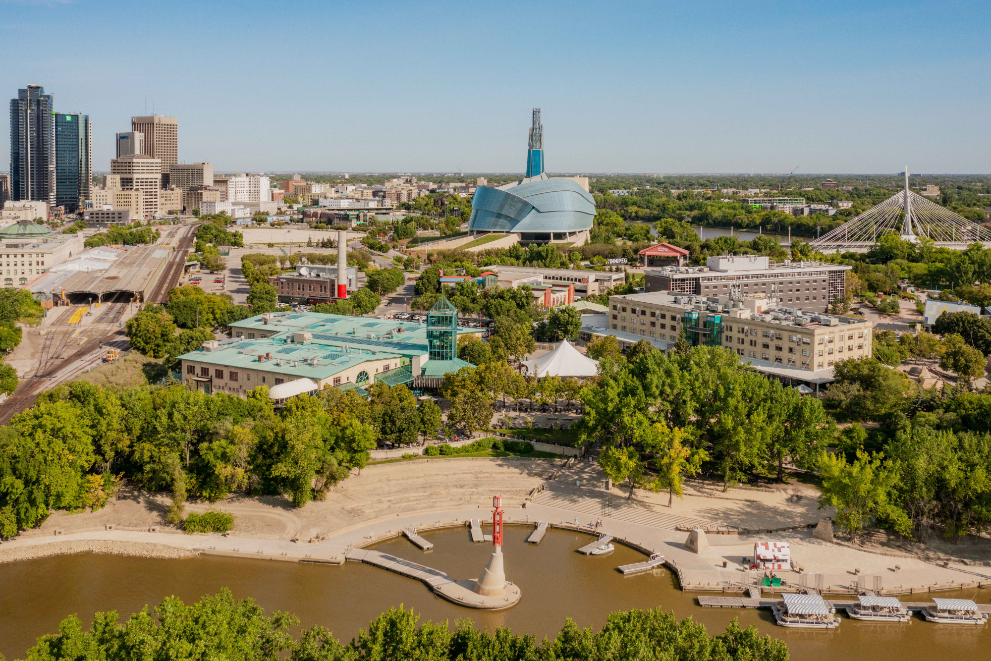 A city skyline view featuring modern skyscrapers, a large contemporary building with a curved roof, and a white cable-stayed bridge over a river, with green trees and a small park area in the foreground.