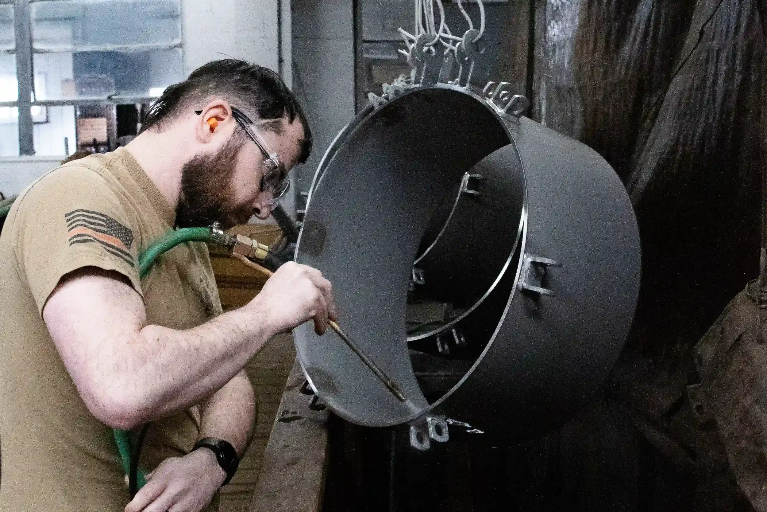 A man working on a large, round metal object in a workshop. He is wearing safety glasses, a tan shirt with an American flag patch, and is focused on welding or assembling the metal piece.