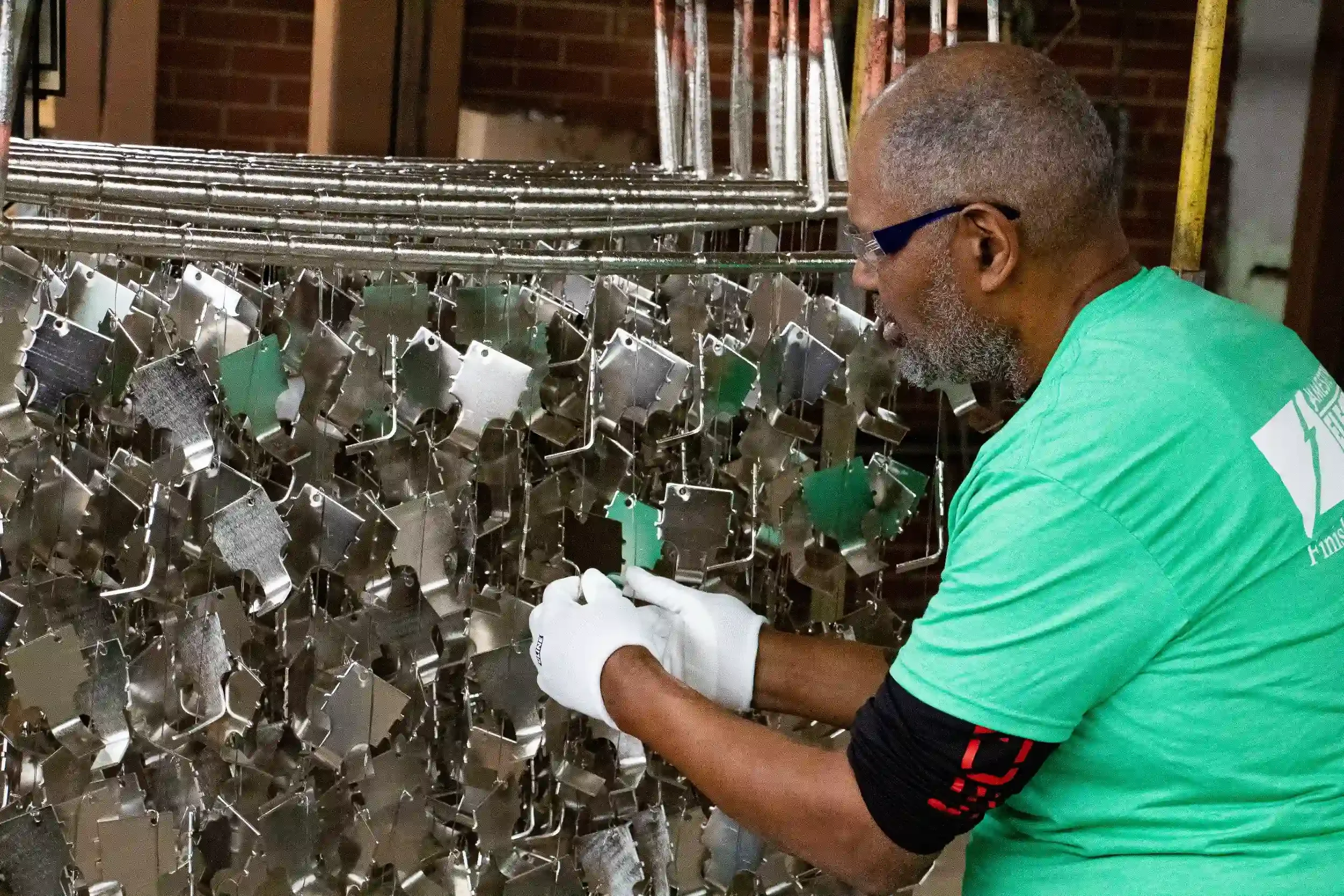 Man with gray beard and glasses inspecting a metal sculpture made of many hanging metal pieces in an industrial setting.