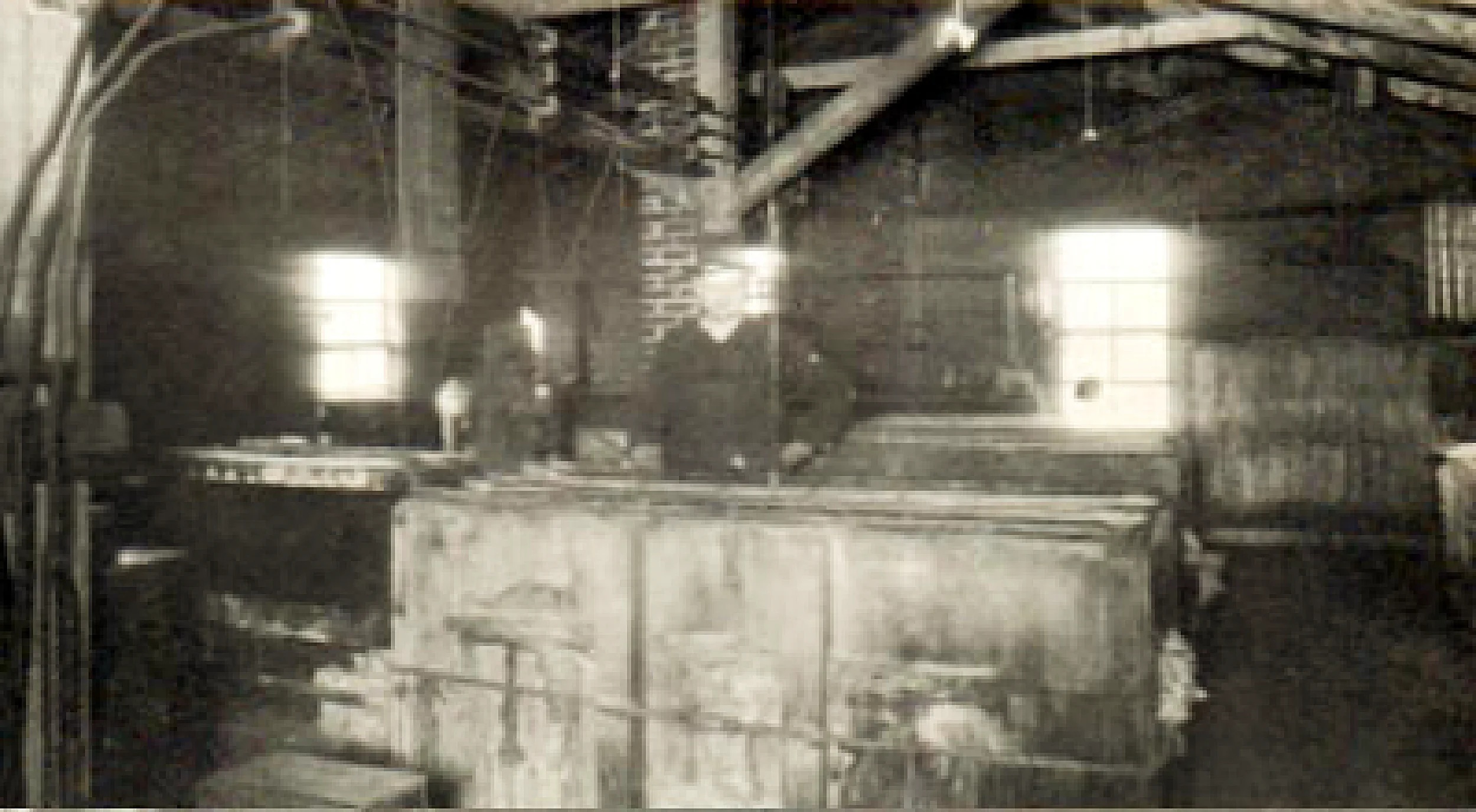 Black and white photo of an industrial building, showing a steel structure, beams, and a staircase, with bright light coming through windows.