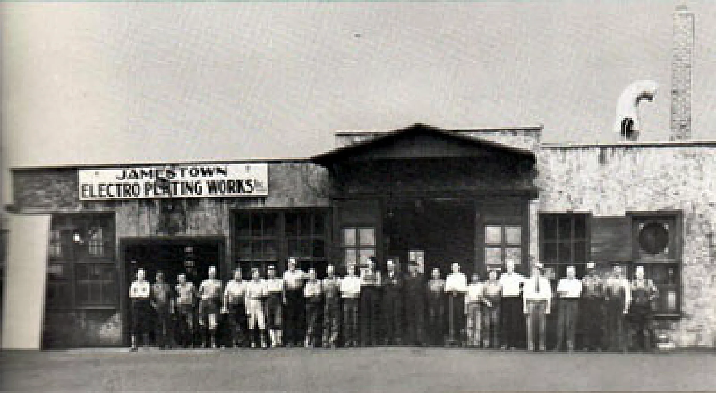 Black and white photo of a group of workers standing in front of a building with a sign that reads 'Jamestown Electro Plating Works.' The workers are wearing uniform shirts and hats, arranged in a single line, facing the camera.