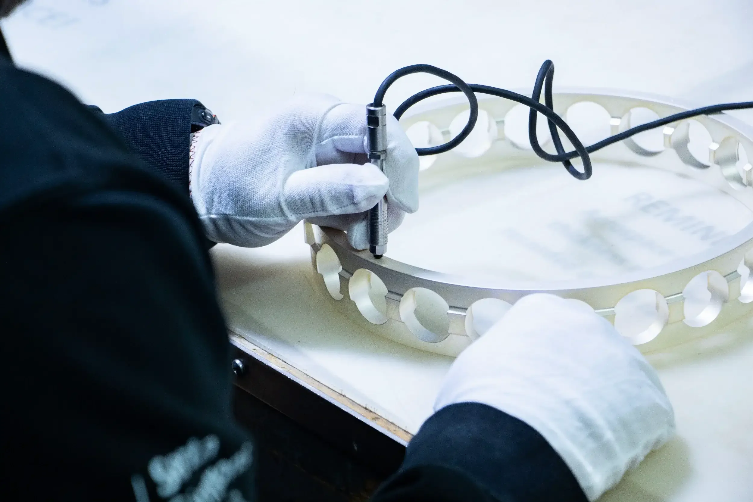 A person wearing a white glove operating a tool on a circular, white metal frame with evenly spaced holes, possibly part of a mechanical or scientific apparatus.