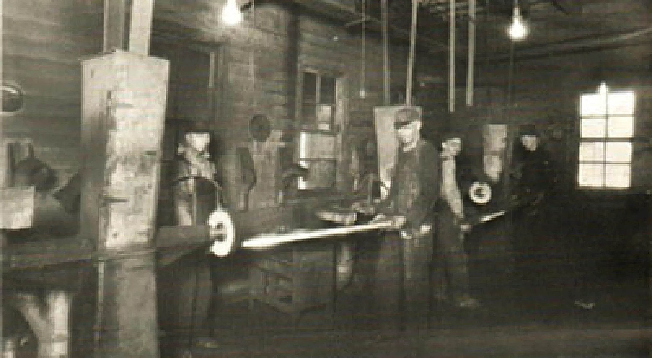 Black and white photo of men working with machinery in an industrial workshop or factory with wooden walls and windows.