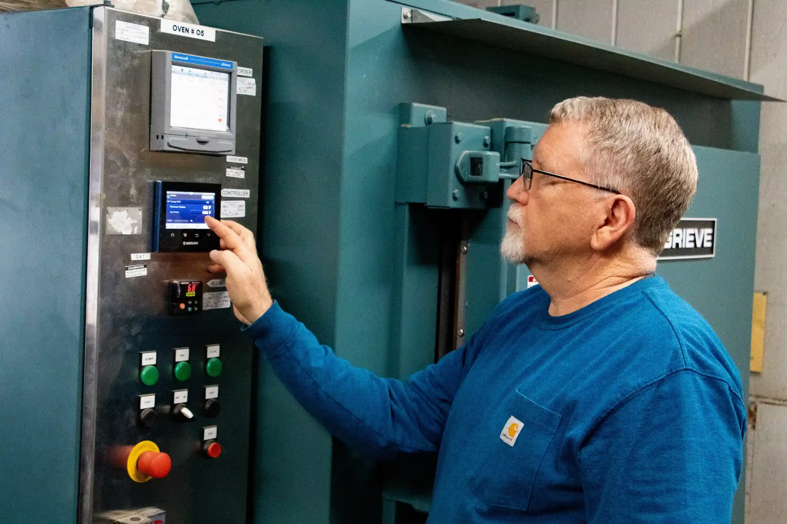 A man wearing glasses and a blue Carhartt shirt is operating a control panel on a large industrial machine in a warehouse or factory setting.