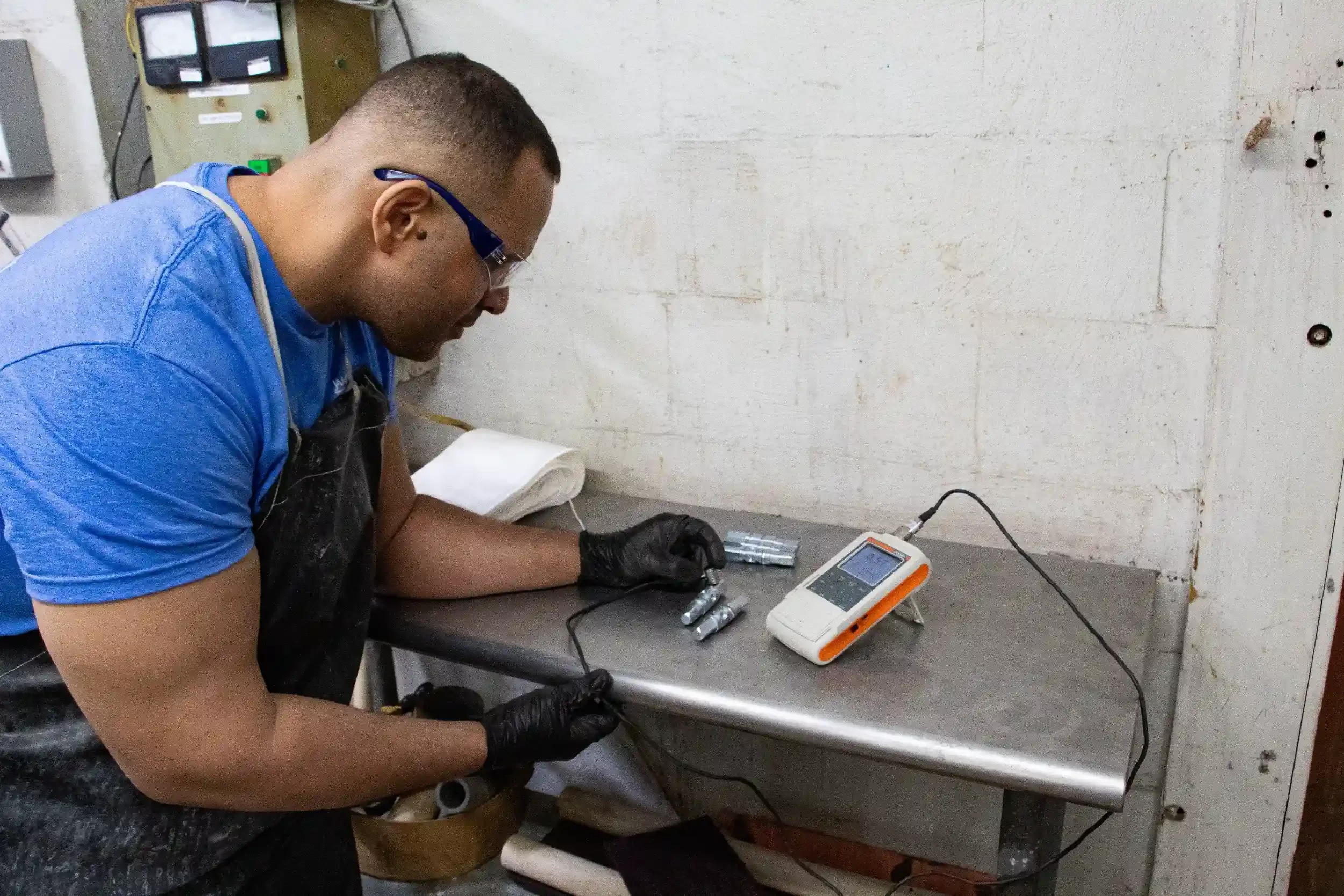 A person wearing glasses and black gloves working with metal parts at a gray workbench, using a testing instrument connected to the parts, in an industrial setting with a white brick wall and electrical equipment in the background.