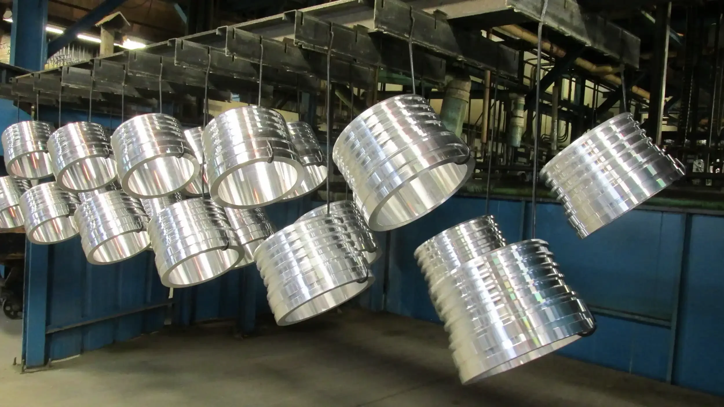 Hanging metal components, possibly parts of a machine or industrial equipment, in a factory setting with industrial pipes and structures in the background.
