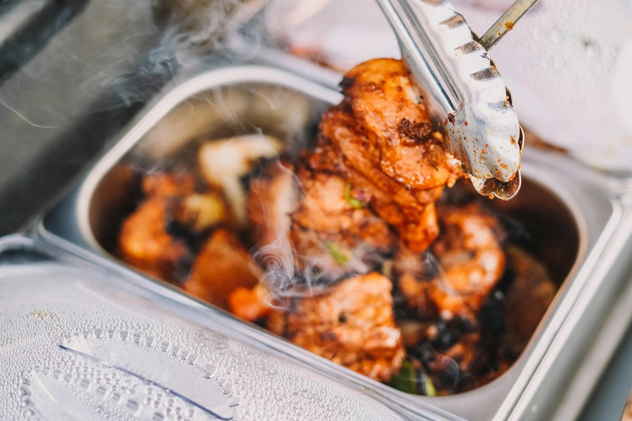 Close-up of a container with cooked marinated chicken kebabs and vegetables, with a meat slicer partially visible on the side.