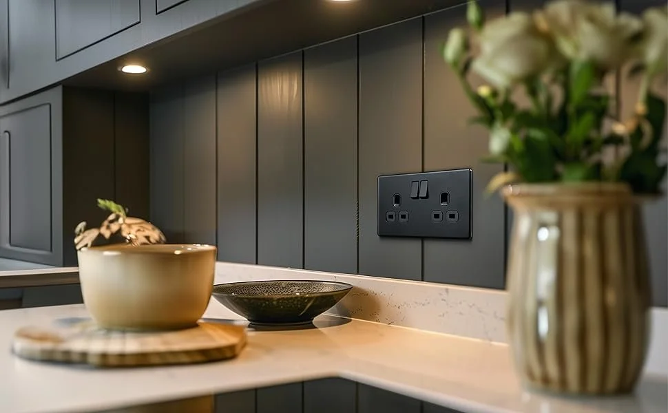 Kitchen countertop with a beige bowl, black plate, and a striped beige and brown vase with white flowers, with dark gray cabinets and a black electrical outlet in the background.