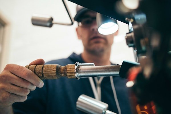 A person wearing glasses and a hoodie working on a metal piece using a handheld tool, with a focused expression and a magnifying light.
