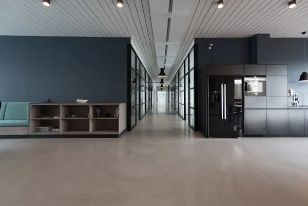 Modern office corridor with glass walls, a teal sofa with a wooden shelf, and black kitchen appliances on the right.