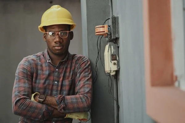 A man with glasses, wearing a yellow hard hat and a plaid shirt, standing with his arms crossed next to an electrical box on a gray wall