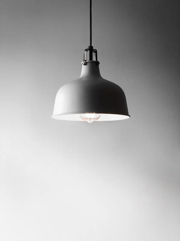 A black pendant ceiling light fixture hanging from a cord, turned on, against a plain, light gray background.
