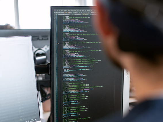 Person with glasses looking at a computer monitor displaying lines of colored code in an office.