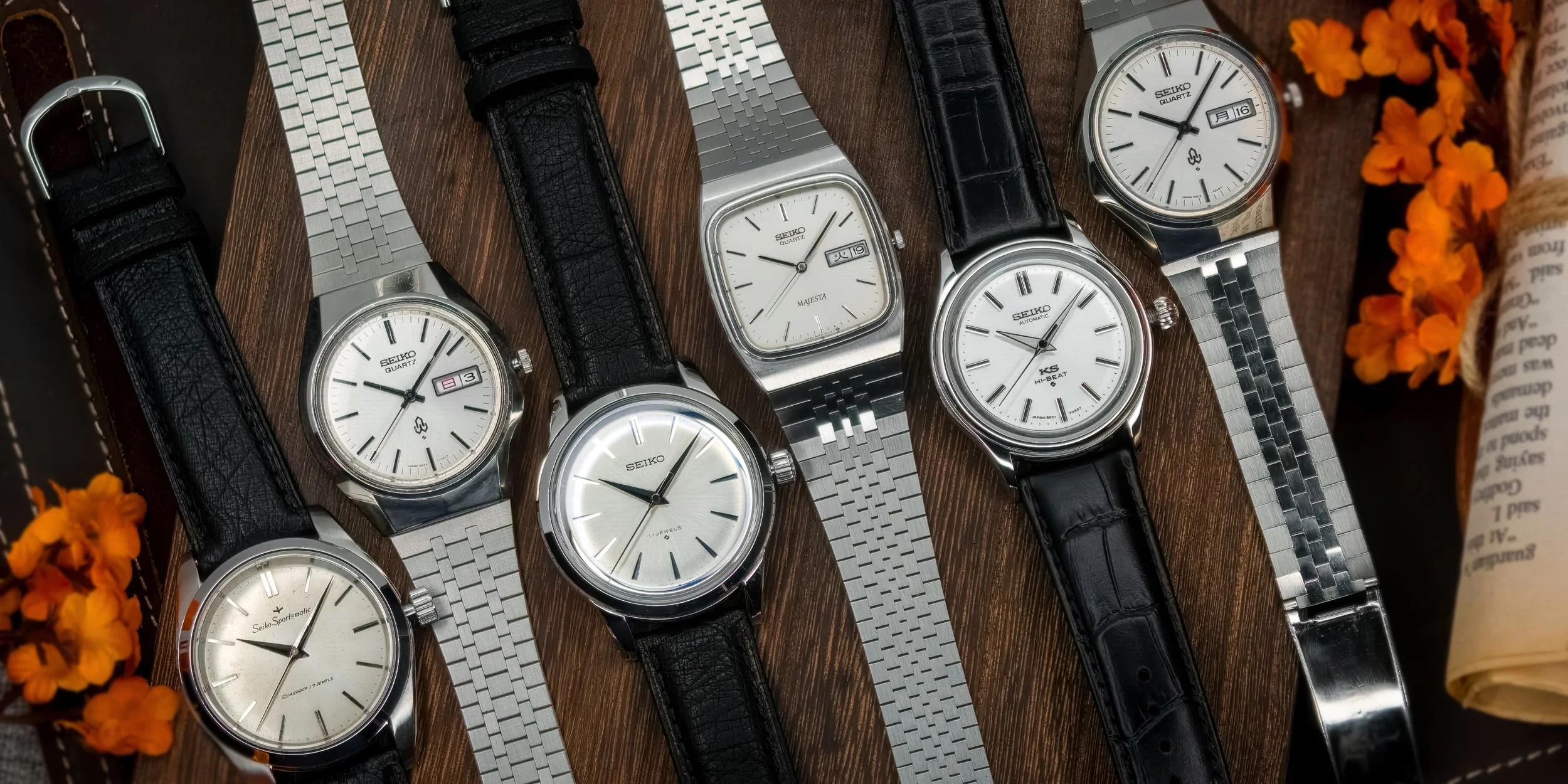 Ulike Seiko-klokker med svarte lærreimer og metallreimer, plassert på et trebord, omgitt av blomster og en avis.