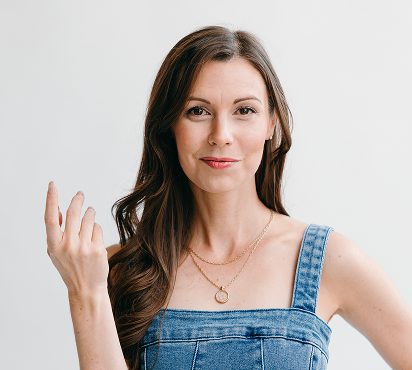 A woman with long brown hair wearing a denim dress, making a gesture with her hand against a plain white background.