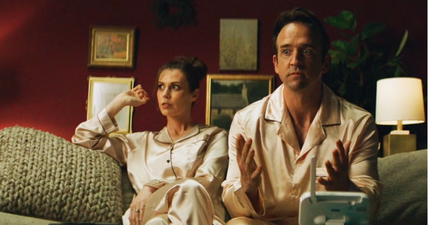 A woman and a man sitting on a sofa in pajamas, with the woman flexing her arm. The man has an animated expression and is gesturing with his hands, in a cozy room with framed pictures, a lamp, and a large plant.