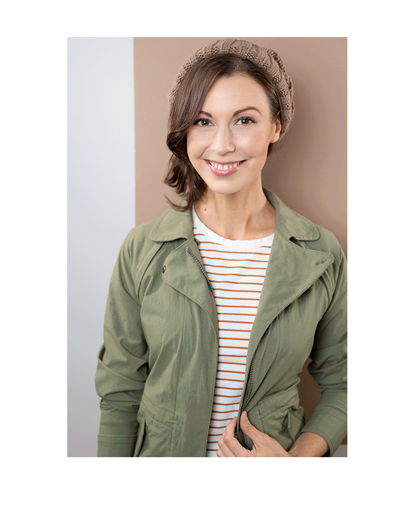 A young woman with shoulder-length brown hair, smiling, wearing a beige knitted beret, an olive green jacket, and a white and red striped shirt, standing against a background with beige and gray walls.