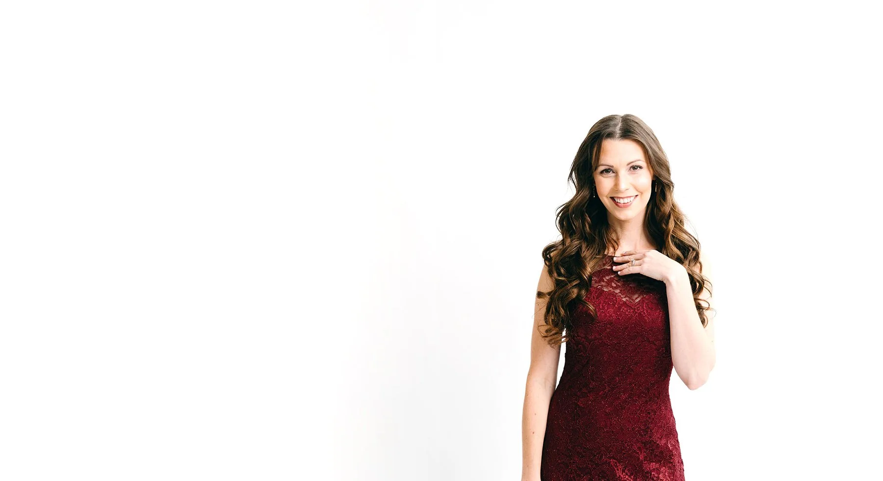 A woman with long brown curly hair wearing a red lace sleeveless dress, smiling, with her left hand gently touching her chest, against a white background.