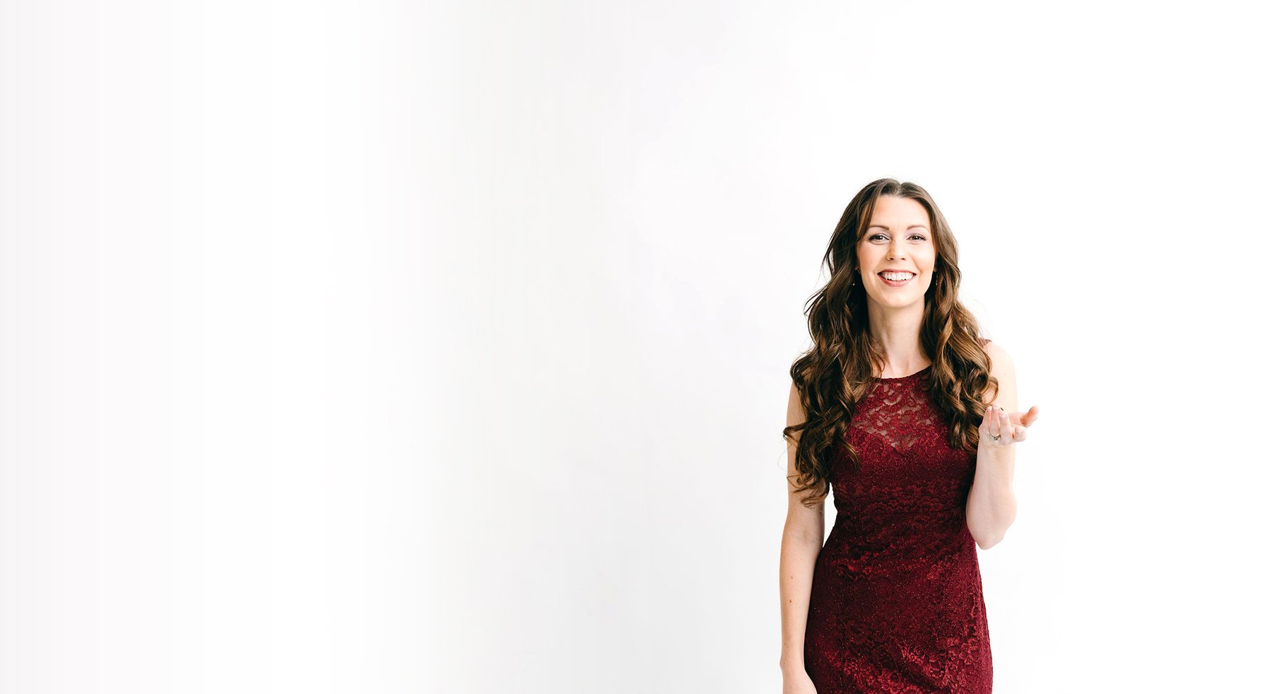 A woman with long, curly brown hair wearing a sleeveless red lace dress smiling and gesturing with her right hand against a white background.