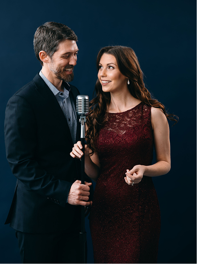 A man and woman standing close together, smiling, with the man holding a vintage microphone between them, against a dark blue background.